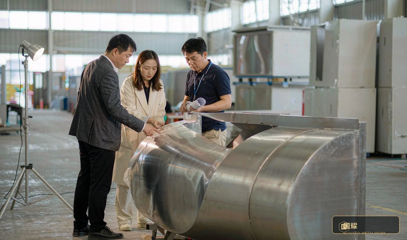 Designer, client and factory project manager reviewing a full-size curved stainless steel mock-up under a raking inspection lamp
