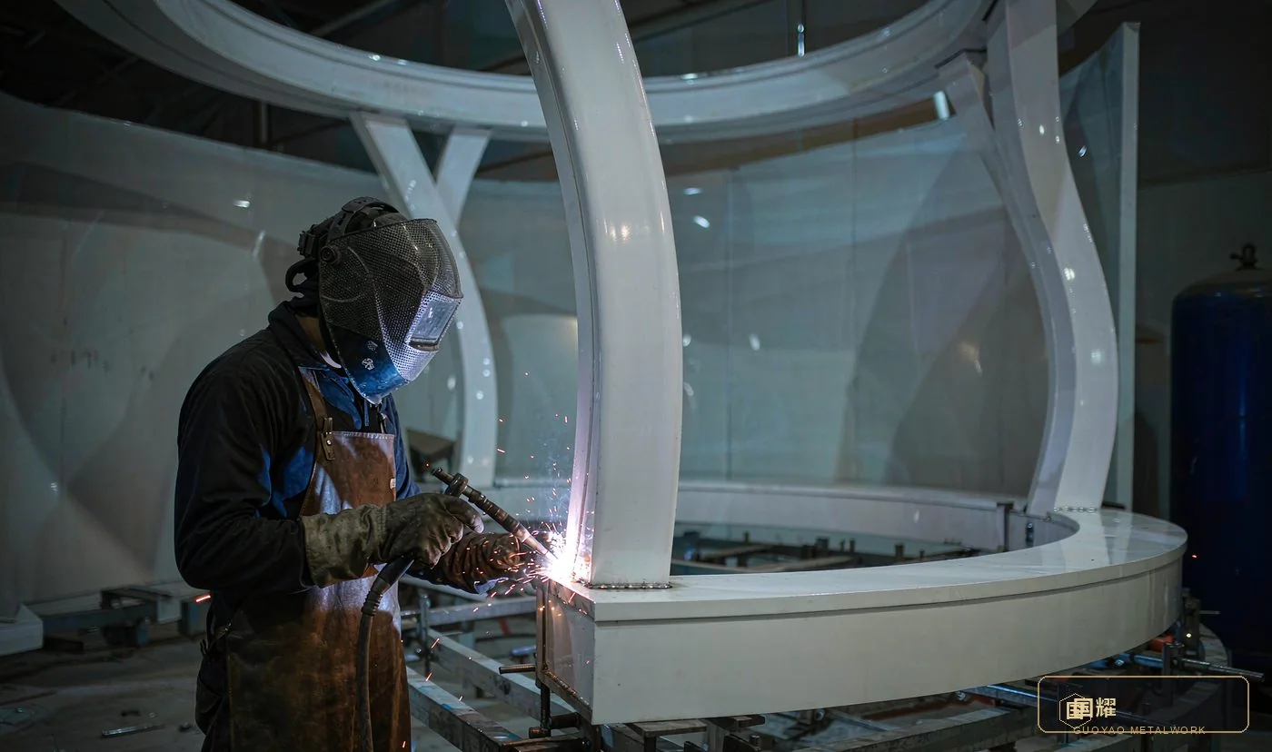 Welder joining a curved white column to an oval ring beam on a jig